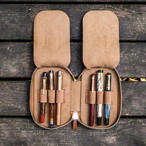 May include: A brown leather pen case with six slots for pens. The case is open and has several pens inside, including a black pen, a brown pen, a gold pen, a red pen, a black and white pen, and a blue pen.