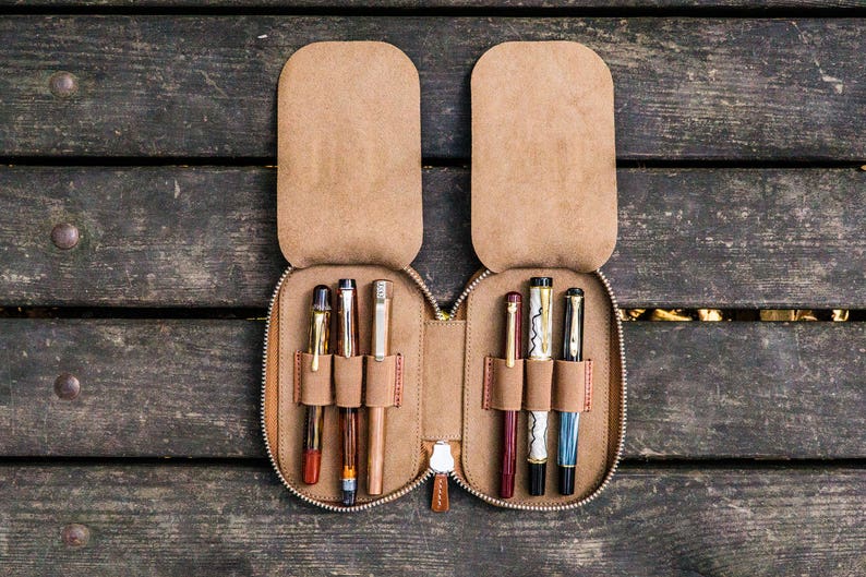 May include: A brown leather pen case with six slots for pens. The case is open and has several pens inside, including a black pen, a brown pen, a gold pen, a red pen, a black and white pen, and a blue pen.