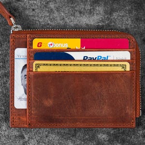 May include: A brown leather wallet with a zipper closure and multiple card slots. The wallet is filled with various credit cards and an ID card. The zipper pull is brown leather. The wallet is on a gray surface.