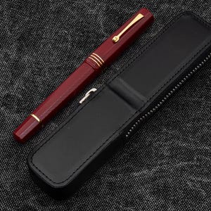 Op de afbeelding: Een bordeauxrode vulpen met goudkleurige accenten en een zwart leren etui met een rits. De etui is open en de pen ligt ernaast op een gestructureerd grijs oppervlak.
