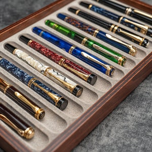 May include: A collection of luxury fountain pens in a wooden display case. The pens feature various colors, including blue, green, and brown, with gold-colored accents. The case has a dark wood finish and a gray interior.