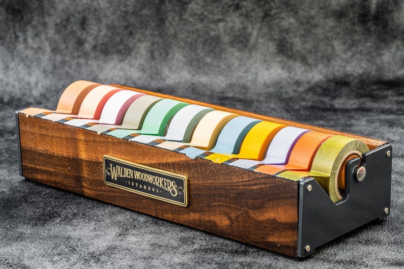 May include: A wooden tape dispenser with a black metal base and a label that reads "Walden Woodworkers - Istanbul". The dispenser holds eight rolls of colorful tape.