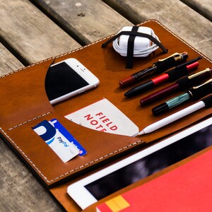 May include: A brown leather portfolio with white stitching detail. The portfolio has multiple pockets and slots for holding a phone, cards, pens, and a tablet. The portfolio is open and a red notebook is visible inside.