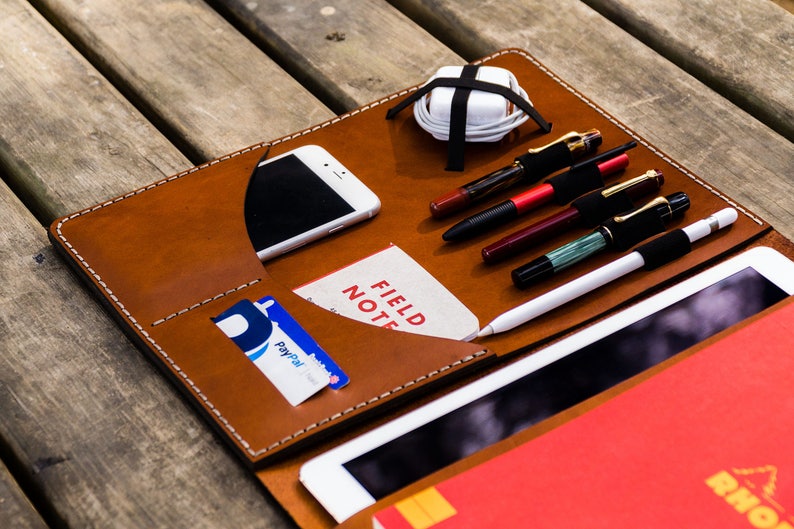 May include: A brown leather portfolio with white stitching detail. The portfolio has multiple pockets and slots for holding a phone, cards, pens, and a tablet. The portfolio is open and a red notebook is visible inside.