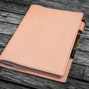 Puede incluir: Un cuaderno de cuero rosa claro con un portaplumas y un bolígrafo insertado. El cuaderno está cerrado y sobre una superficie de madera oscura.