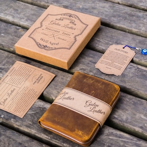 May include: A brown leather journal with a zipper closure and a brown leather strap with the text "Galen Leather" printed on it. The journal is on a wooden surface with a small brown tag that says "Our Story" and a blue evil eye charm.