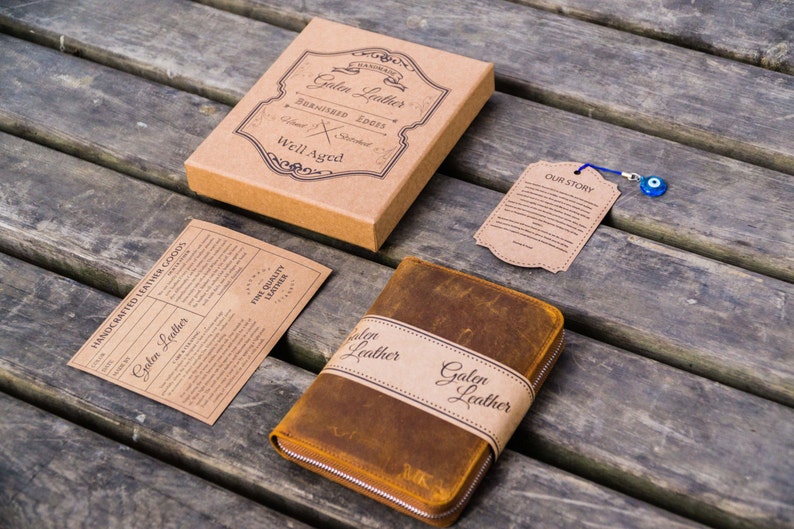 May include: A brown leather journal with a zipper closure and a brown leather strap with the text "Galen Leather" printed on it. The journal is on a wooden surface with a small brown tag that says "Our Story" and a blue evil eye charm.