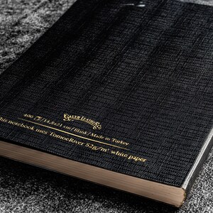 K&ouml;nnte beinhalten: Ein schwarzes Notizbuch mit goldener Beschriftung, die "Gallery Leather Co." und "This notebook uses Tomoe River 52g/m&sup3; white paper 400/14,821 cm/tilink/Made in Turkey" lautet.