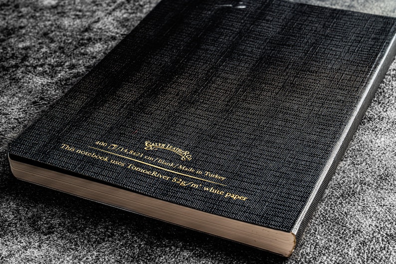 K&ouml;nnte beinhalten: Ein schwarzes Notizbuch mit goldener Beschriftung, die "Gallery Leather Co." und "This notebook uses Tomoe River 52g/m&sup3; white paper 400/14,821 cm/tilink/Made in Turkey" lautet.