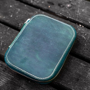May include: A teal leather zippered pouch with white stitching. The pouch is lying on a dark wooden surface.
