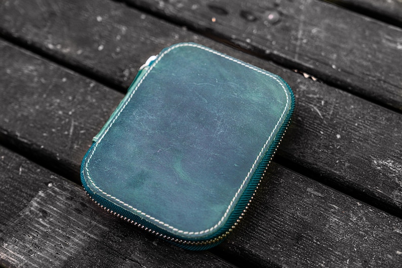 May include: A teal leather zippered pouch with white stitching. The pouch is lying on a dark wooden surface.