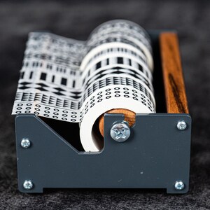 May include: A grey and wood tape dispenser with a white patterned tape roll. The tape has a black and white geometric design.