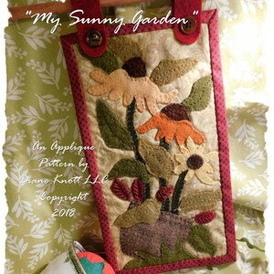 Könnte beinhalten: Ein handgefertigter Filz-Applikations-Wandbehang mit dem Titel "My Sunny Garden". Das Design zeigt eine Vase mit Blumen in Orange-, Gelb- und Grüntönen. Der Text "An Applique Pattern by Diane Knott LLC Copyright 2018" ist unterhalb des Designs sichtbar.