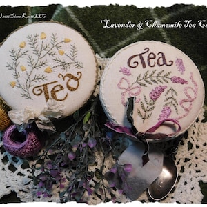 May include: Two round tea caddies with embroidered floral designs and the word "Tea". One caddy features yellow flowers, the other lavender. A tea bag and spoon are included. The caddies are on a white lace doily.