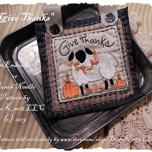 Może przedstawiać: Wzór do haftu lub haftu igłowego przedstawiający białą owcę z czarnymi oczami i kopytami, otoczoną jesiennymi liśćmi i dyniami. Wzór nosi tytuł "Give Thanks" i jest autorstwa Diane Knett LLC.