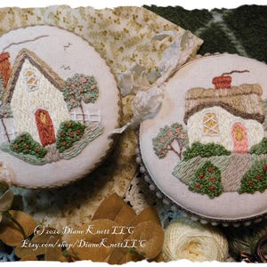 May include: Two round embroidered cottage scenes. Each features a white house with a thatched roof, a red or pink door, and surrounding trees and bushes. The embroidery is on a cream-colored fabric with a scalloped edge.