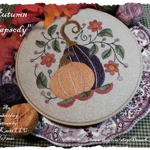 Op de afbeelding: Een borduurpatroon met de titel "Herfst Rhapsody" met een paarse en oranje pompoen met groene bladeren en roze bloemen. Het patroon wordt getoond in een hoepel op een tafel met garen en een schaar.