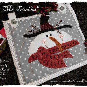 May include: A grey and white fabric applique pattern featuring a snowman with a black hat, red scarf, and a brown nose. The pattern is titled "Mr. Twinkles" and is by Diane Knott LLC.
