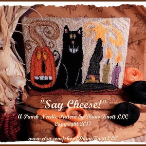 May include: A punch needle pattern for a Halloween-themed pillow featuring a black cat, a pumpkin, and candles. The pattern is titled "Say Cheese!" and is by Diane Knott LLC. Copyright 2017.