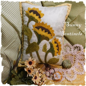 May include: A white pillow with an applique design of three yellow sunflowers with green stems and leaves. The pillow is surrounded by a green and white gingham fabric, a crocheted doily, and a small bouquet of yellow daisies. The text "Sunny Sentinels" is written in cursive on the pillow.