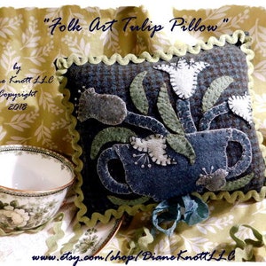 May include: A decorative pillow with a folk art tulip design. The pillow features a dark blue background with appliqued white tulips, green leaves, and a blue cup. The pillow is trimmed with a green, wavy border. The text "Folk Art Tulip Pillow" is at the top.