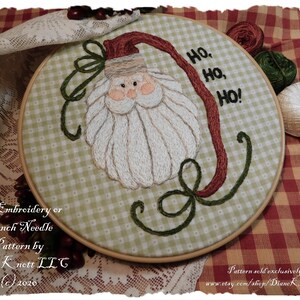 May include: An embroidery or punch needle pattern featuring Santa Claus with the text "Ho, Ho, Ho!" on a green and white checkered background. The design is in a wooden hoop, with the artist's name and copyright information at the bottom.
