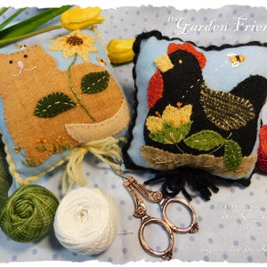 Puede incluir: Dos alfileteros de fieltro, uno con un diseño de gato y girasol y el otro con una gallina negra y flores amarillas. Los alfileteros están sobre un fondo azul con lunares blancos. El texto "Garden Friends" está en la parte superior de la imagen.