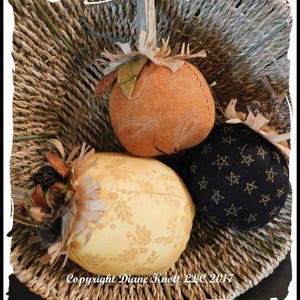 May include: Three fabric pumpkins in a woven basket. One pumpkin is orange with a white design, one is yellow with a brown design, and one is black with white stars.