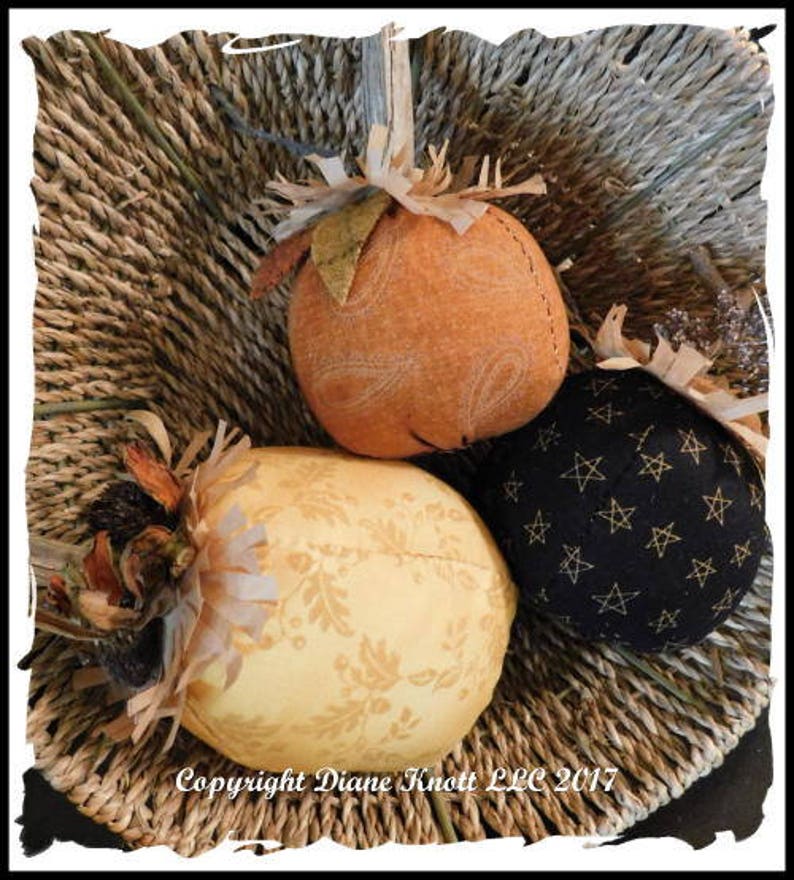May include: Three fabric pumpkins in a woven basket. One pumpkin is orange with a white design, one is yellow with a brown design, and one is black with white stars.