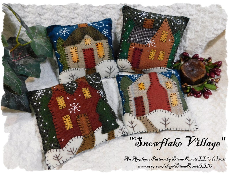 May include: Four felt applique pillow ornaments with a winter village theme. Each pillow features a different house design with a red roof, white snow, and green trees. The pillows are decorated with snowflakes and a white background. The text "Snowflake Village" is written in cursive at the bottom of the image.
