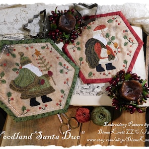 May include: Two hexagon-shaped embroidered Christmas ornaments featuring Santa Claus. One ornament depicts Santa carrying a sack of gifts, the other depicts Santa holding a Christmas tree. The ornaments are surrounded by red berries and green foliage. The text "Woodland Santa Duo" is visible at the bottom of the image.