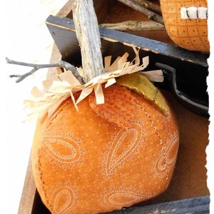 May include: A handmade orange pumpkin with a paisley pattern, a twig stem, and brown paper shreds. The pumpkin is sitting in a wooden box with a black handle.