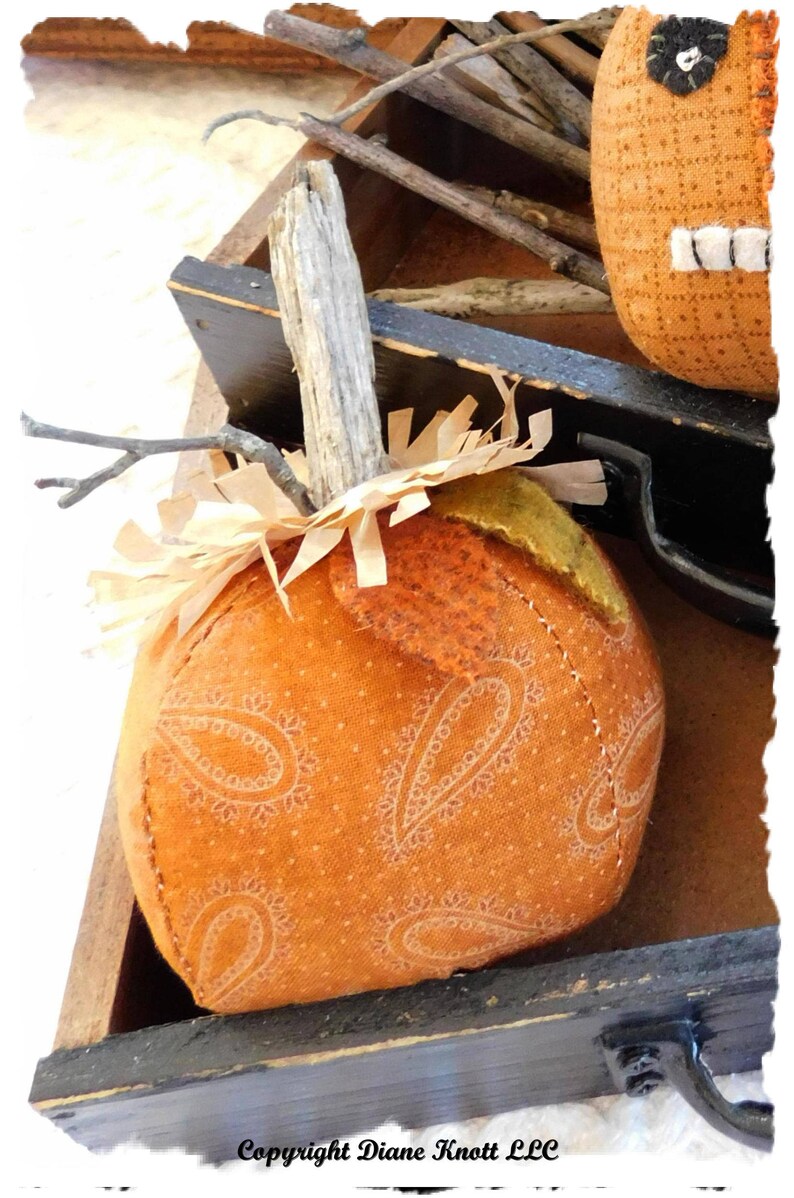 May include: A handmade orange pumpkin with a paisley pattern, a twig stem, and brown paper shreds. The pumpkin is sitting in a wooden box with a black handle.