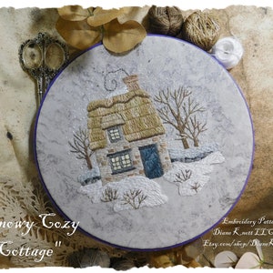 Puede incluir: Diseño bordado de una acogedora cabaña con techo de paja, rodeada de nieve y árboles. El diseño está en un trozo de tela redonda en un aro. El texto "Snowy Cozy Cottage" está escrito debajo del aro.