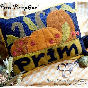 May include: A decorative pillow with a navy blue background and a yellow border. The pillow features appliqued orange pumpkins, green leaves, and the word "Prim" in large letters. The text "Prim Pumpkins" is at the top.