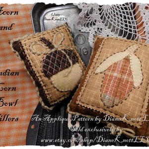 May include: Two brown fabric applique patterns, one with an acorn design and the other with an Indian corn design. The patterns are titled "Acorn and Indian Corn Bowl Fillers" and are an applique pattern by Diane Knott LLC.