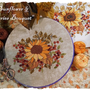 Może przedstawiać: Haftowany wzór słonecznika i bukietu jagód na beżowym materiale z fioletowym wykończeniem. Wzór otoczony jest brązowymi, pomarańczowymi i beżowymi kulkami włóczki oraz parą antycznych nożyczek. Tekst "Sunflower & Berries Bouquet" jest napisany kursywą nad wzorem.