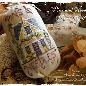 May include: A beige needle roll with embroidered text and a detailed house design. The text reads "PINS & NEEDLES" and "Needle Roll". The house is yellow with a gray roof and purple door, surrounded by floral embroidery. A decorative item for sewing.
