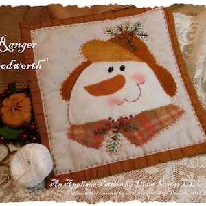May include: A brown and white applique pattern of a snowman wearing a brown hat and scarf with a sprig of greenery. The pattern is titled "Ranger Woodworth" and is by Diane K nett LLC.