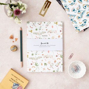 May include: A white notebook with a floral pattern in shades of pink, green, and brown. The notebook says "NOTES" and "Sarah W" on the front cover. The notebook is on a light pink surface with a pencil, a pencil sharpener, and a small book.