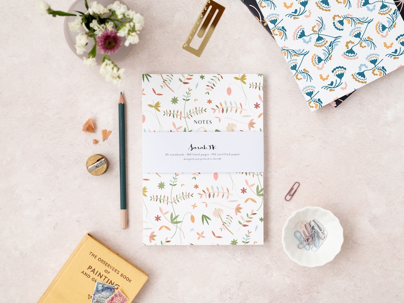May include: A white notebook with a floral pattern in shades of pink, green, and brown. The notebook says "NOTES" and "Sarah W" on the front cover. The notebook is on a light pink surface with a pencil, a pencil sharpener, and a small book.