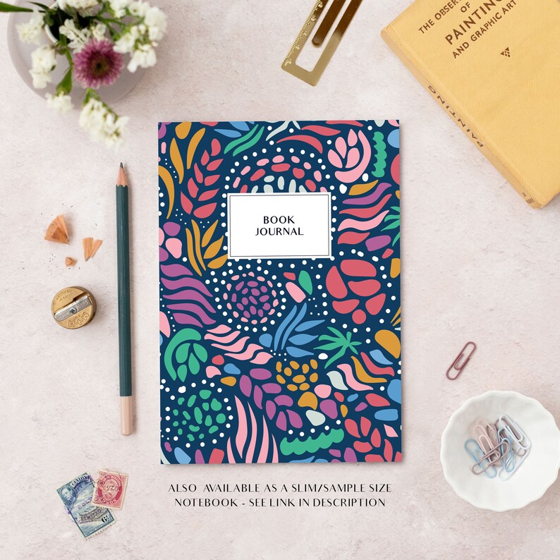 May include: A navy blue journal with a vibrant abstract floral pattern. The cover has a white rectangle with the text "BOOK JOURNAL". A pencil, sharpener, and book are also visible.