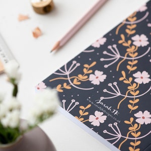 May include: A black notebook with a floral pattern in pink, orange, and white. The notebook has the text "Sarah TK" and "www.sarahjkdesigns.co.uk" printed on the cover.