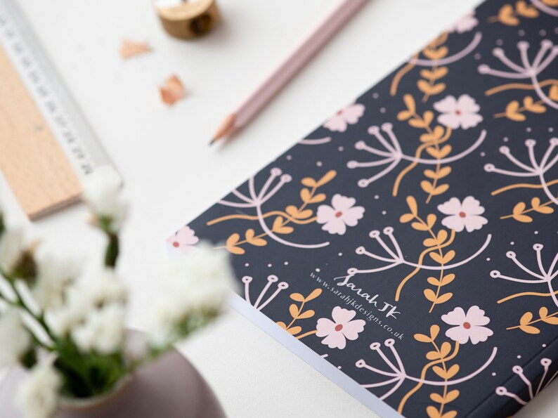 May include: A black notebook with a floral pattern in pink, orange, and white. The notebook has the text "Sarah TK" and "www.sarahjkdesigns.co.uk" printed on the cover.