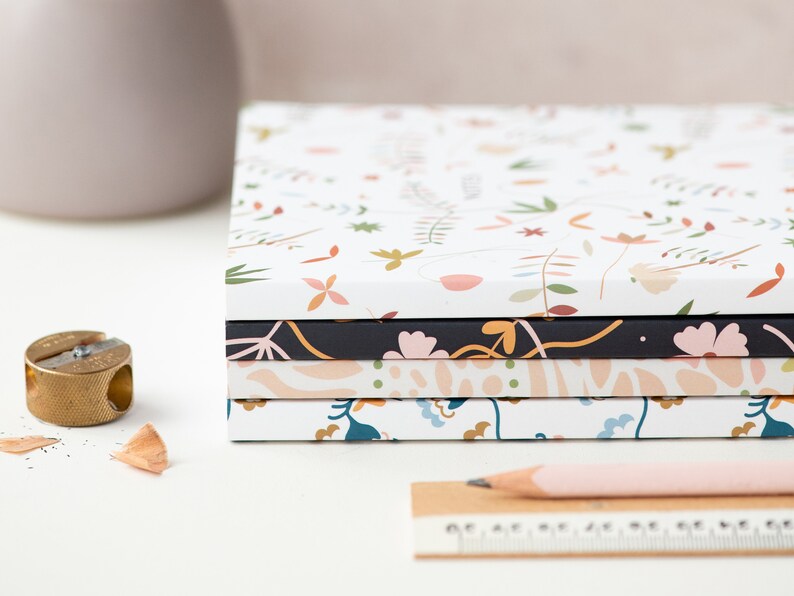 May include: A stack of five notebooks with floral patterns. The top notebook has a white background with small pink, orange, and green flowers. The second notebook has a black background with pink flowers and green leaves. The third notebook has a peach background with abstract shapes. The fourth notebook has a white background with blue and green flowers. The bottom notebook has a white background with small pink, orange, and green flowers.