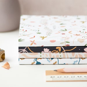 May include: A stack of five notebooks with floral patterns. The top notebook has a white background with small pink, orange, and green flowers. The second notebook has a black background with pink flowers and green leaves. The third notebook has a light pink background with abstract shapes. The fourth notebook has a white background with blue and green flowers. The bottom notebook has a white background with small pink, orange, and green flowers.