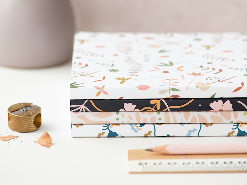 May include: A stack of five notebooks with floral patterns. The top notebook has a white background with small pink, orange, and green flowers. The second notebook has a black background with pink flowers and green leaves. The third notebook has a light pink background with abstract shapes. The fourth notebook has a white background with blue and green flowers. The bottom notebook has a white background with small pink, orange, and green flowers.