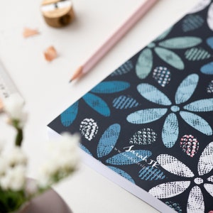 May include: A notebook with a dark blue cover featuring a floral pattern in shades of blue, white, and pink. A pencil, a pencil sharpener, and a wooden ruler are also visible on the white surface. The notebook has the text "Sarah JK Design".
