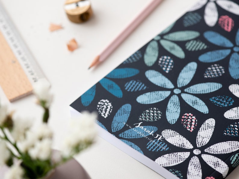 May include: A notebook with a dark blue cover featuring a floral pattern in shades of blue, white, and pink. A pencil, a pencil sharpener, and a wooden ruler are also visible on the white surface. The notebook has the text "Sarah JK Design".