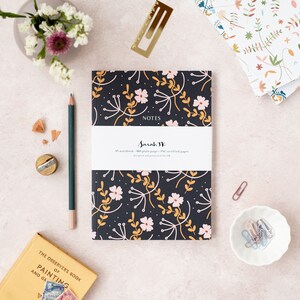 May include: A black notebook with a floral pattern in pink, yellow, and white. The notebook says "NOTES" and "Sarah JK" on the front cover. The notebook is on a light pink surface with a pencil, a pencil sharpener, and a book.